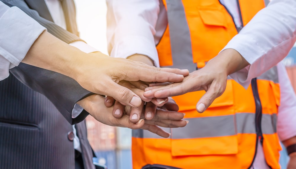 A group of professionals in a teamwork gesture, stacking their hands together in the center. The team consists of individuals in business attire and others wearing high-visibility orange safety vests, symbolizing a collaborative effort between office and field workers. The background is blurred, focusing on the unity and commitment of the team in a workplace safety context.