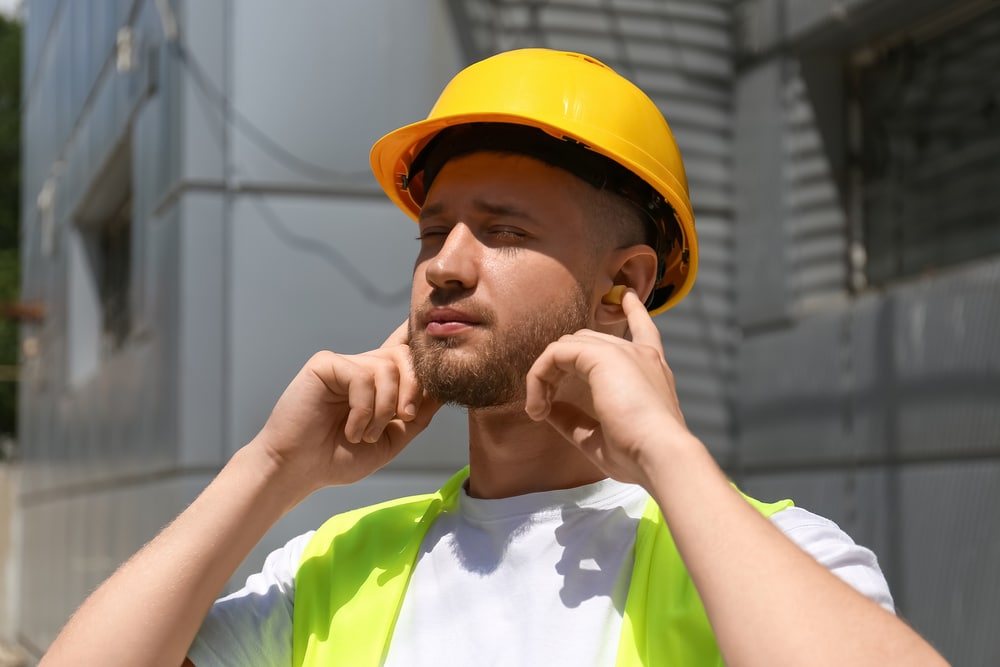 A man wearing a hard hat is inserting ear plugs to protect his hearing from noise at work.