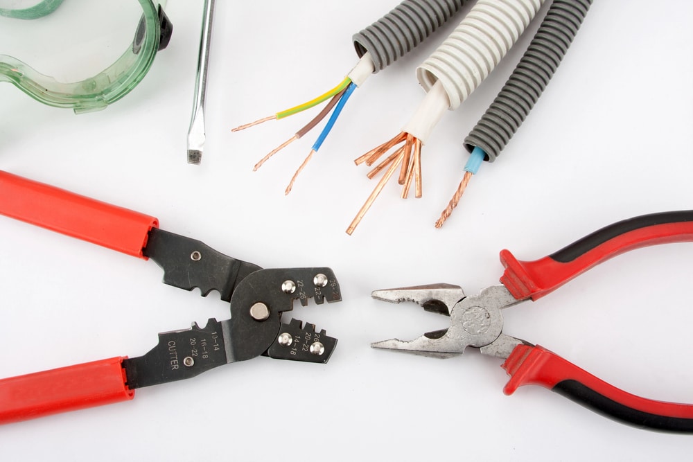 An assortment of electrical tools and components on a white background. In the foreground, there are red-handled wire stripping and crimping tools to the left and combination pliers to the right. Behind these tools are exposed multicolored electrical wires, some encased in white insulation and others in flexible grey conduit. A pair of green safety goggles and a silver hex key lie towards the back. The setup suggests electrical maintenance work.