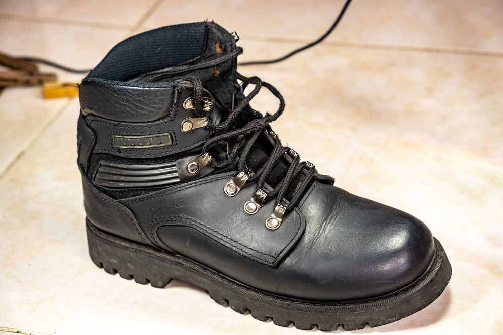 A single black rubber-soled safety boot positioned on a tiled floor. The boot features a steel toe cap, leather upper, and sturdy laces, with metallic eyelets. Visible on the boot are labels that read 'TOP LINE,' indicating the brand. The boot is designed for industrial or electrical work, providing protection and support for the foot with additional electric shock protection.