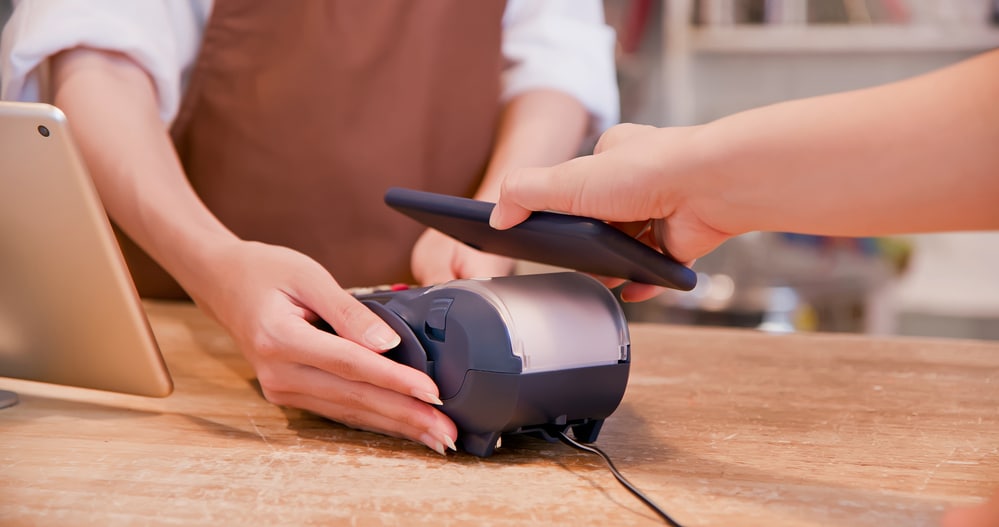 Business customer making a mobile payment using NFC technology at a point of sale terminal with a tablet in the background. They are purchasing personal protective equipment for their employees.