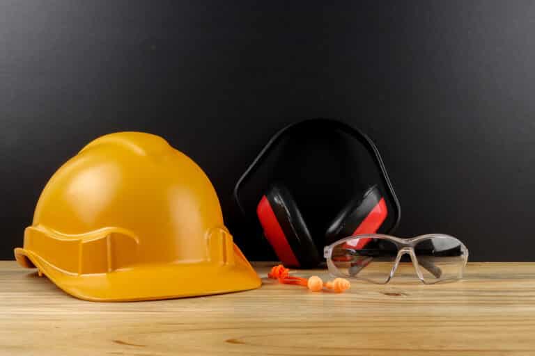 Essential personal protective equipment on a wooden workbench against a black background, featuring a yellow hard hat, red and black ear defenders, clear safety goggles, and orange earplugs.
