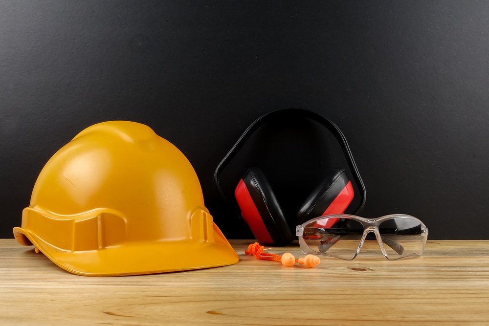Essential personal protective equipment on a wooden workbench against a black background, featuring a yellow hard hat, red and black ear defenders, clear safety goggles, and orange earplugs.