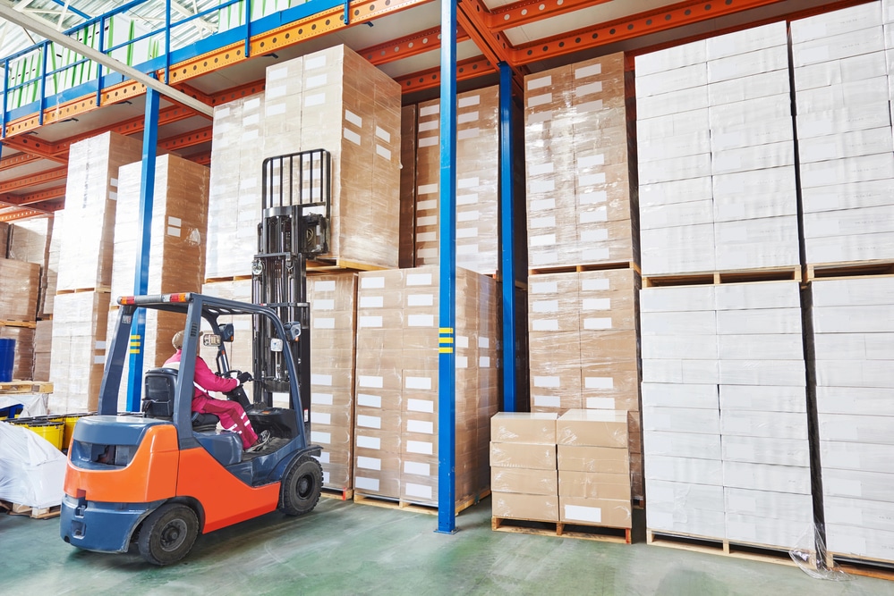 A warehouse worker operates a forklift to lift pallets stacked with cardboard boxes. The scene is inside a large warehouse with high shelves filled to capacity with similar pallets. Natural light illuminates the space from windows near the roof.