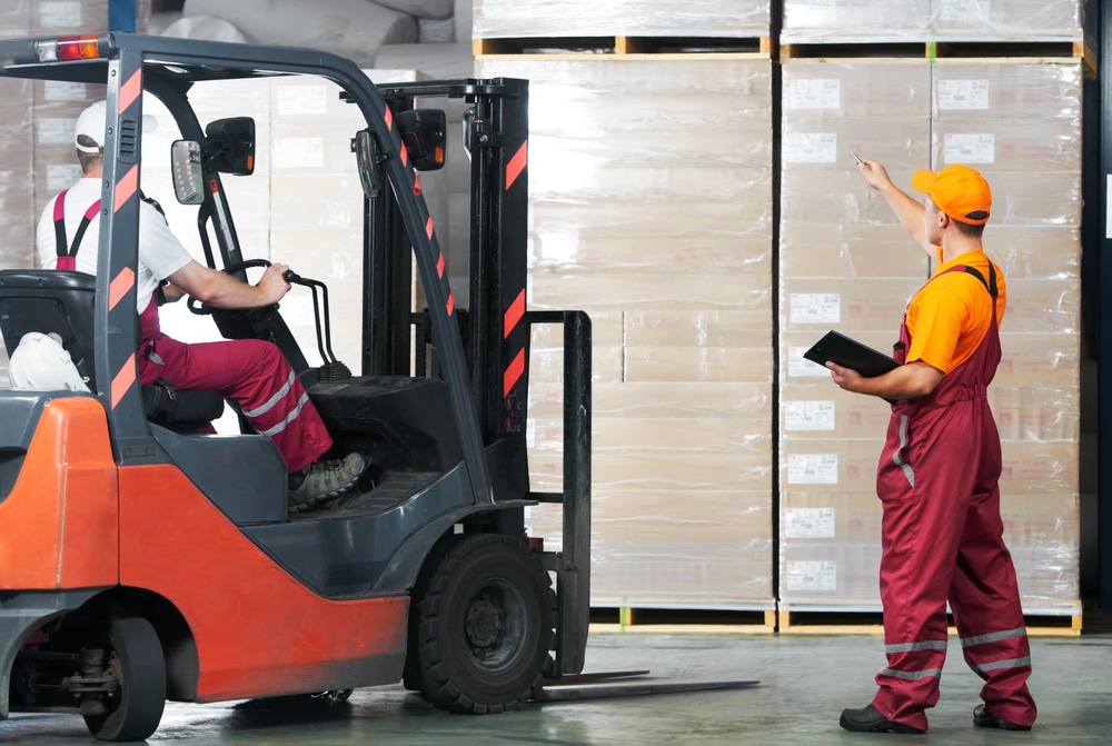 Inside a well-lit warehouse, two workers in safety gear are coordinating the movement of goods. One worker, seated in an orange and black forklift, looks forward while operating the vehicle. Another worker, standing nearby wearing an orange cap, maroon jumpsuit, and holding a digital tablet, gestures towards the high shelves filled with wrapped pallets, seemingly directing or counting inventory.