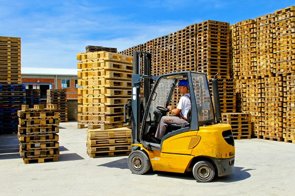 A worker operates a yellow forklift in an outdoor pallet storage area under clear blue skies. The forklift is moving between rows of stacked wooden pallets, which are arranged in tall, organized stacks. The background features a warehouse with visible windows, emphasizing an industrial setting.