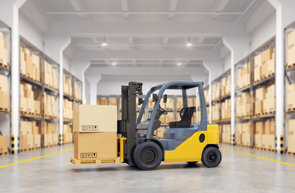 A yellow forklift stands idle in the middle of a spacious, modern warehouse. The forklift is loaded with a pallet carrying multiple cardboard boxes, ready for transport. The background shows neatly organized aisles with numerous shelves stocked with similar boxes, all under a well-lit ceiling.