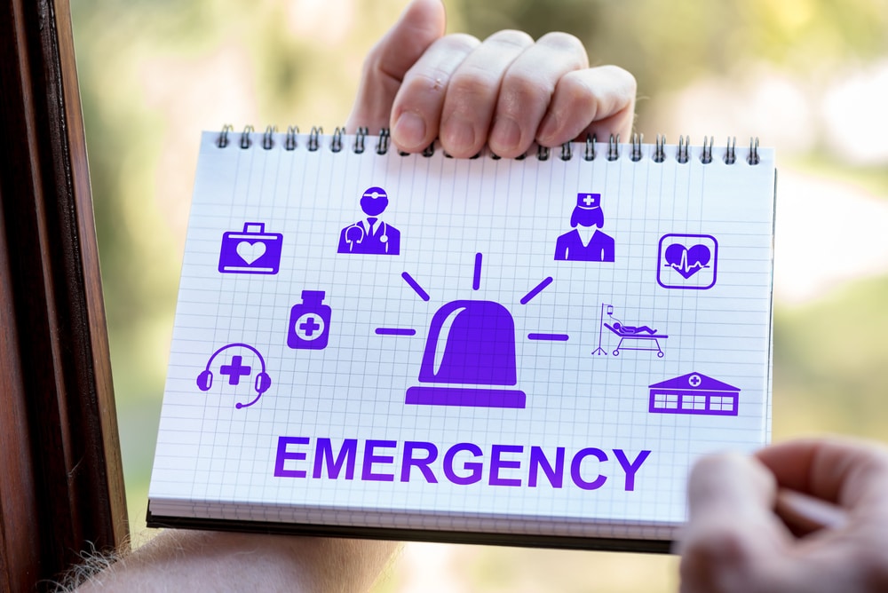 Notebook with emergency symbols being held by a person. The notebook displays various icons representing emergency services such as a doctor, nurse, medical bag, ambulance, hospital building, and an emergency alarm. The word 'EMERGENCY' is prominently written at the bottom. The image is intended to convey the importance of emergency preparedness and response.