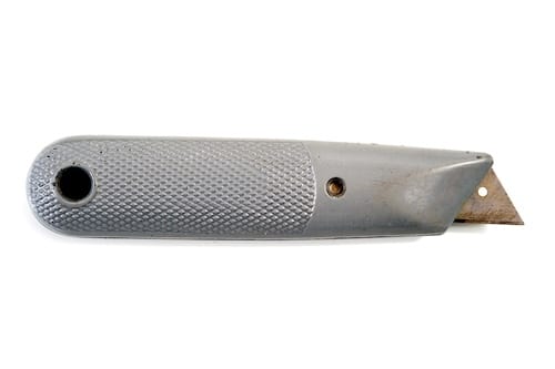 The image shows a silver-colored box cutter with a textured grip. The box cutter has a retractable blade extending slightly from the angled metal casing. The handle has a knurled pattern for better grip, and there is a circular hole near the base, possibly for attaching a lanyard or for hanging. The tool appears to be well-used, with some wear visible on the surface.