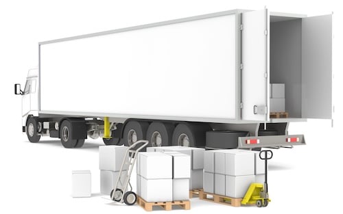 The image shows a white semi-truck with its trailer doors open, being loaded with cargo. Several large cardboard boxes are stacked on wooden pallets near the rear of the trailer. A hand truck and a pallet jack are positioned nearby, indicating that the loading process is either ongoing or about to begin. The trailer is empty except for a few boxes already inside, and the scene appears to be taking place in a warehouse or loading dock area. The truck and equipment are clean and modern, suggesting an efficient, organized operation.
