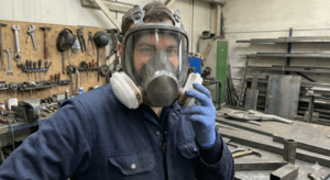 A worker in a metal fabrication shop wearing a 3M full-face air-purifying respirator with dual cartridge filters and blue nitrile gloves, holding an additional filter cartridge.