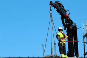 Construction worker in a hi-vis vest and safety harness guides a crane lifting a steel load with a chain sling rigging on a job site.