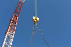 Crawler crane hook block with wire rope slings lifting a load against a clear blue sky—heavy lifting and rigging on a construction site.